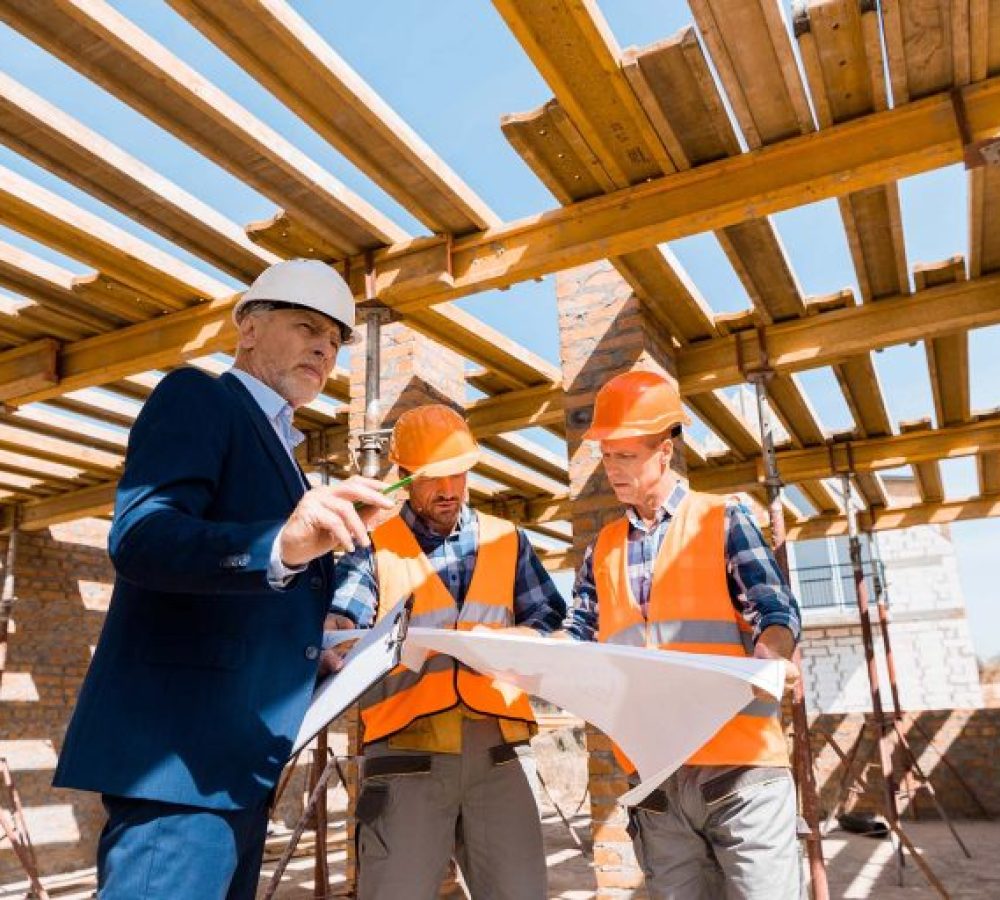 mature-businessman-near-builders-in-helmets-holdin-S6S29SR-768x512